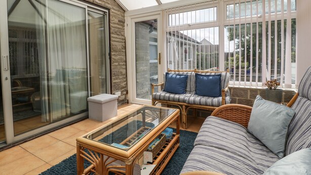 A sunlit patio with a glass door leading to a garden, a wicker sofa with cushions, and a glass coffee table. - Melita (Langport)