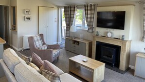 A living room with a fireplace, sofa, armchair, television, and a window with curtains. - Yorkshire View Lodge (York)