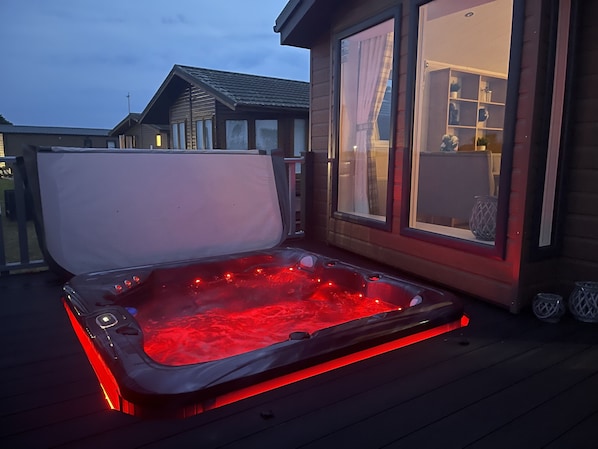 Un jacuzzi iluminado con luz roja, una terraza de madera y un edificio con amplios ventanales.