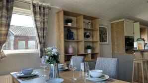 A dining area with a wooden table set for a meal, a window with curtains, and a wooden shelving unit with decorative items. - Yorkshire View Lodge (York)