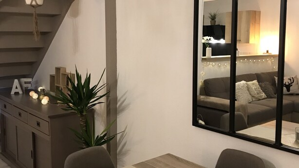 Un comedor moderno con una mesa de madera, un vaso de bebida, velas y un espejo grande que refleja un espacio de sala acogedor con un sofá y una planta.