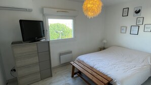 Un dormitorio con cama, un banco de madera, cómoda, televisor, ventana y lámpara de pared.