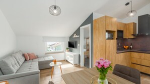 Uma sala moderna com um sofá cinzento, uma mesa de jantar de madeira com um vaso de flores, e uma cozinha com armários de madeira e um micro-ondas preto.