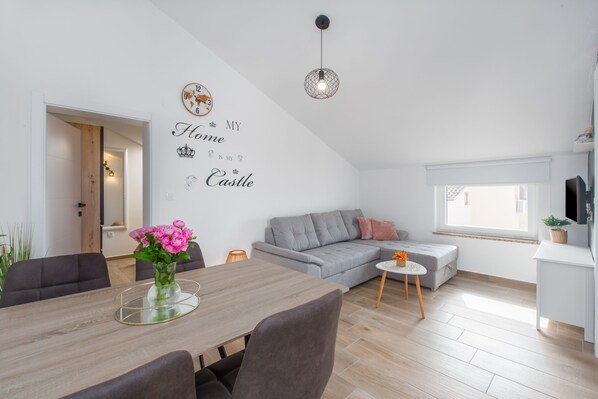A modern living room with a dining area, a grey sofa, a small table with a vase of flowers, and a wall with decorative text. - 250 m from the beach, modern, air-conditioned, sleeps 2 to 3 people (Porec)