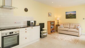 A modern kitchen with white cabinets, a built-in oven, and a microwave. There is a beige sofa and a small table with a lamp in the living area. - Long Batch Cottage (Shepton Mallet)
