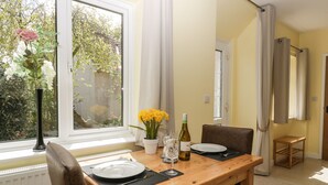 A dining area with a wooden table set for two, a vase with flowers, and a bottle of wine on the windowsill. - Long Batch Cottage (Shepton Mallet)