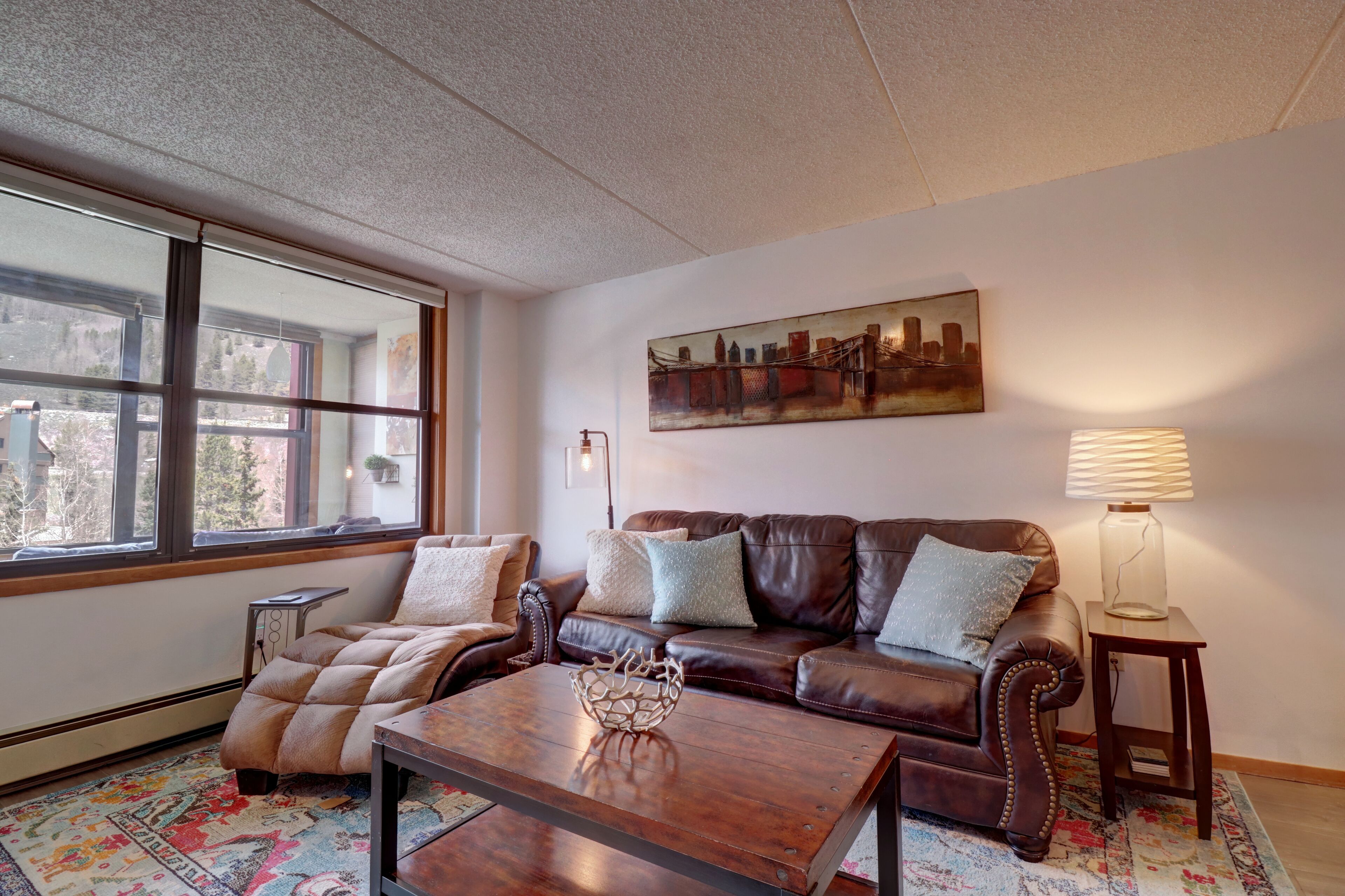 A living room with a brown leather sofa, a wooden coffee table, and a large window with a cityscape view.