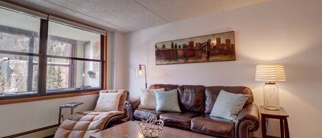 A living room with a brown leather sofa, a wooden coffee table, and a large window with a cityscape view.