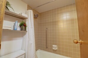 Een badkamer met een bad, een douche en een toilet. Er zijn planken met toiletartikelen en een potplant.