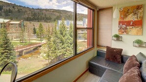 A room with a large window offering a view of a bridge, trees, and buildings. There is a black leather sofa with two cushions and a painting on the wall.