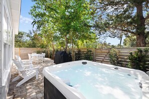 A hot tub in a backyard with a wooden fence, white chairs, and trees.