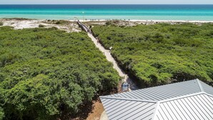 Luftaufnahme einer Küstenlandschaft mit Strand, einem Wanderweg durch dichtes Grün und einem Gebäude mit Metallbedachung.