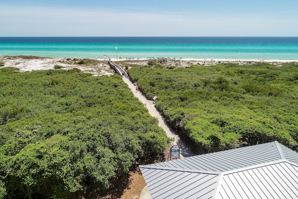 Vista aérea de una zona costera con playa, un sendero para peatones a través de vegetación densa y una estructura con techo de metal.