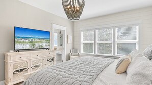 Ein Schlafzimmer mit einem großen Bett, einem Fernseher auf einer Kommode, einem Sessel und einem Fenster mit Jalousien.
