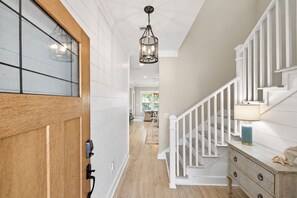 A hallway with a wooden door, a hanging light fixture, a staircase with white railings, and a wooden dresser with drawers. - ☀️ GOLF CART, HEATED POOL, Private Hot Tub, Bikes, Ping-Pong, Walk to Rosemary (Inlet Beach)