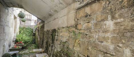 Un estrecho callejón con una pared de piedra, dos sillas de metal y una pequeña zona ajardinada con plantas en macetas.