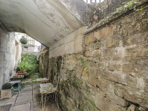 A narrow alleyway with a stone wall, two metal chairs, and a small garden area with potted plants. - Dollar Ward House Basement (Cirencester)