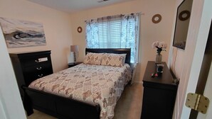 Un dormitorio con cama, mesita de noche, cómoda, lámpara, ventana con cortinas y un cuadro en la pared.