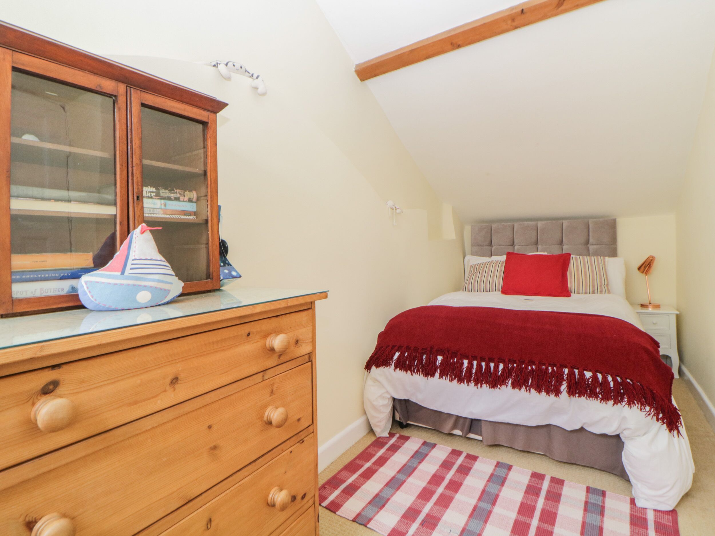 A bedroom with a wooden dresser, a bed with a red blanket, a checkered rug, and a window with a view of the outside.