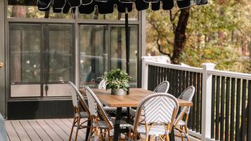 A wooden deck with a table and chairs under a black umbrella, surrounded by a white railing and trees in the background.
