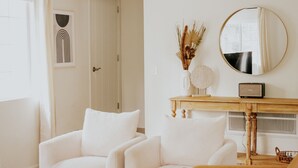 Una sala de estar con dos sillones blancos, una mesa de centro de madera y un sofá marrón. Un espejo redondo cuelga en la pared sobre un mueble bajo de madera con un jarrón de plantas secas.