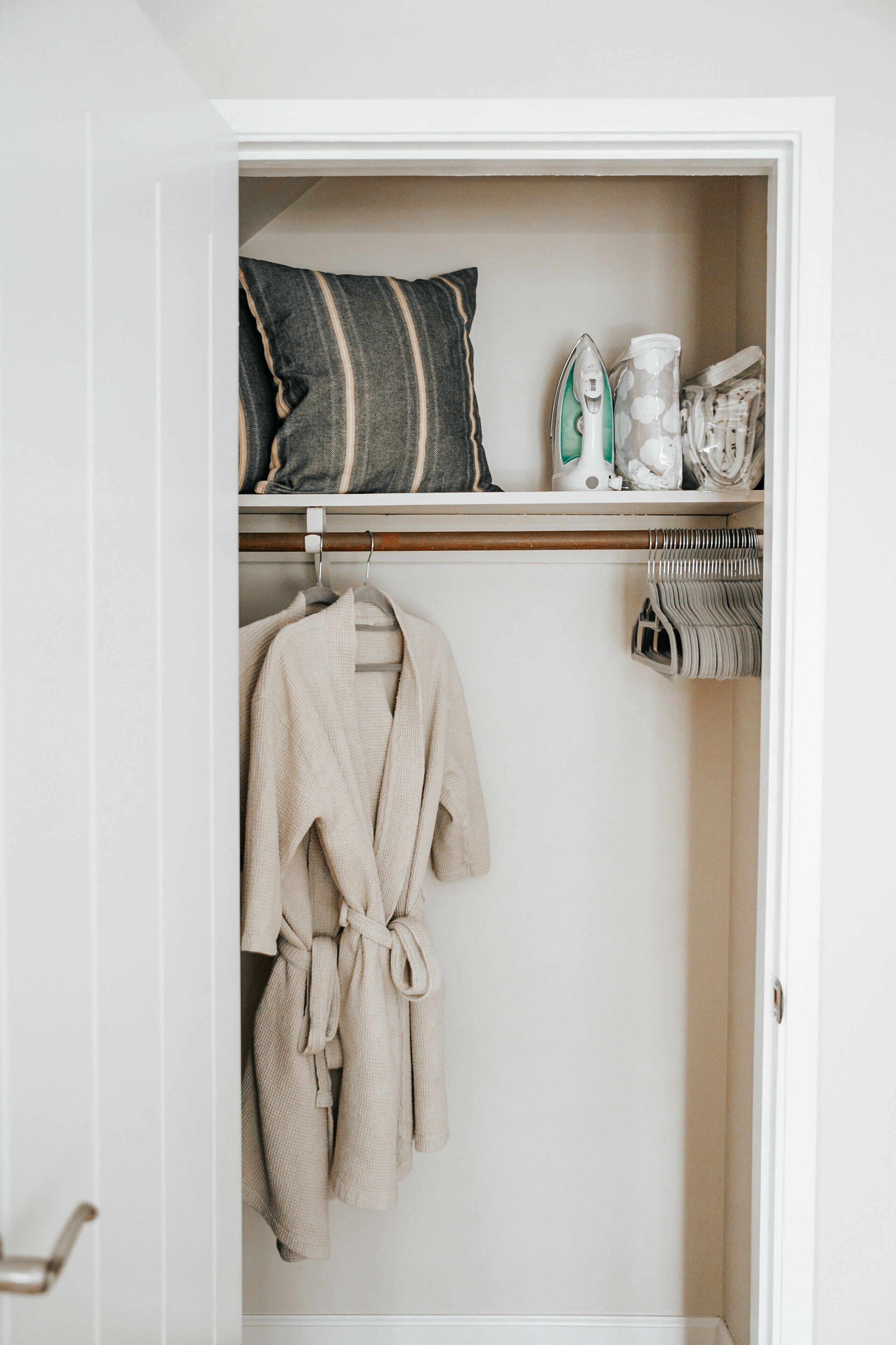 A closet with a robe hanging, a shelf with a pillow, an iron, and a basket.