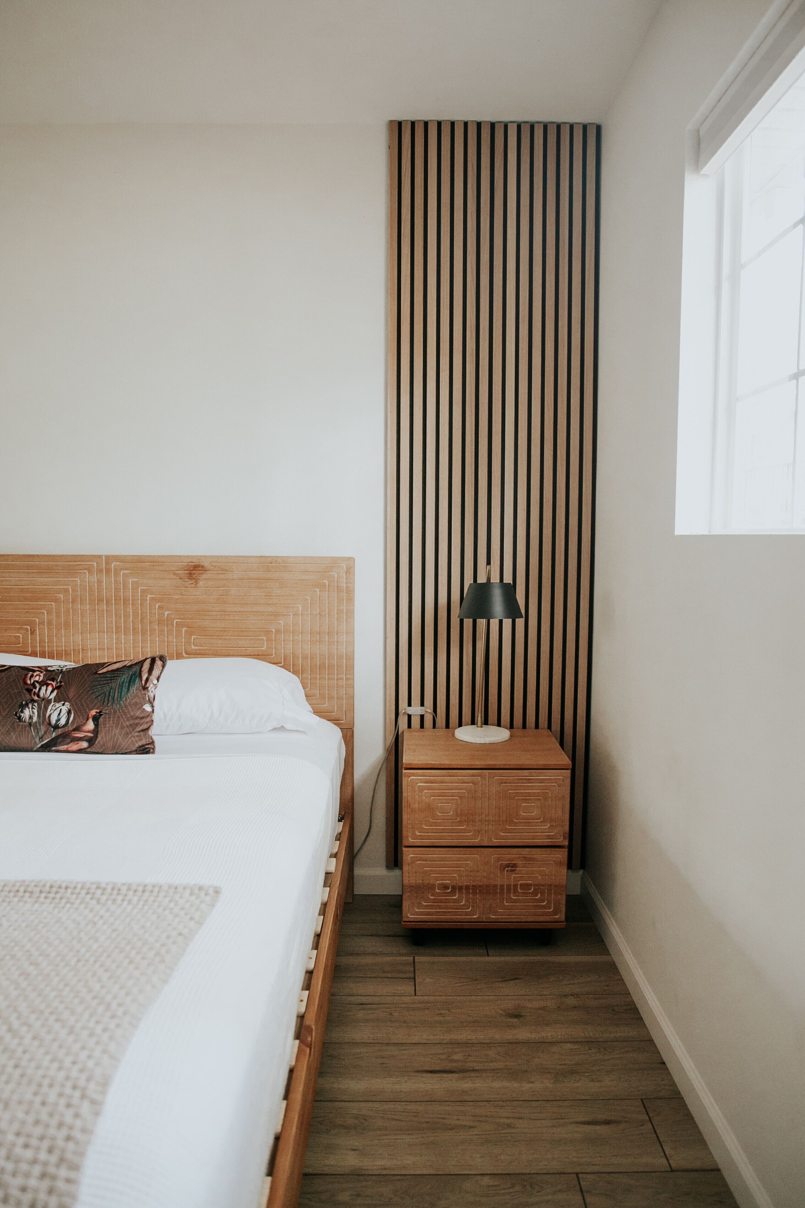 A bedroom with a wooden headboard, a bedside table with a lamp, and a window with curtains.