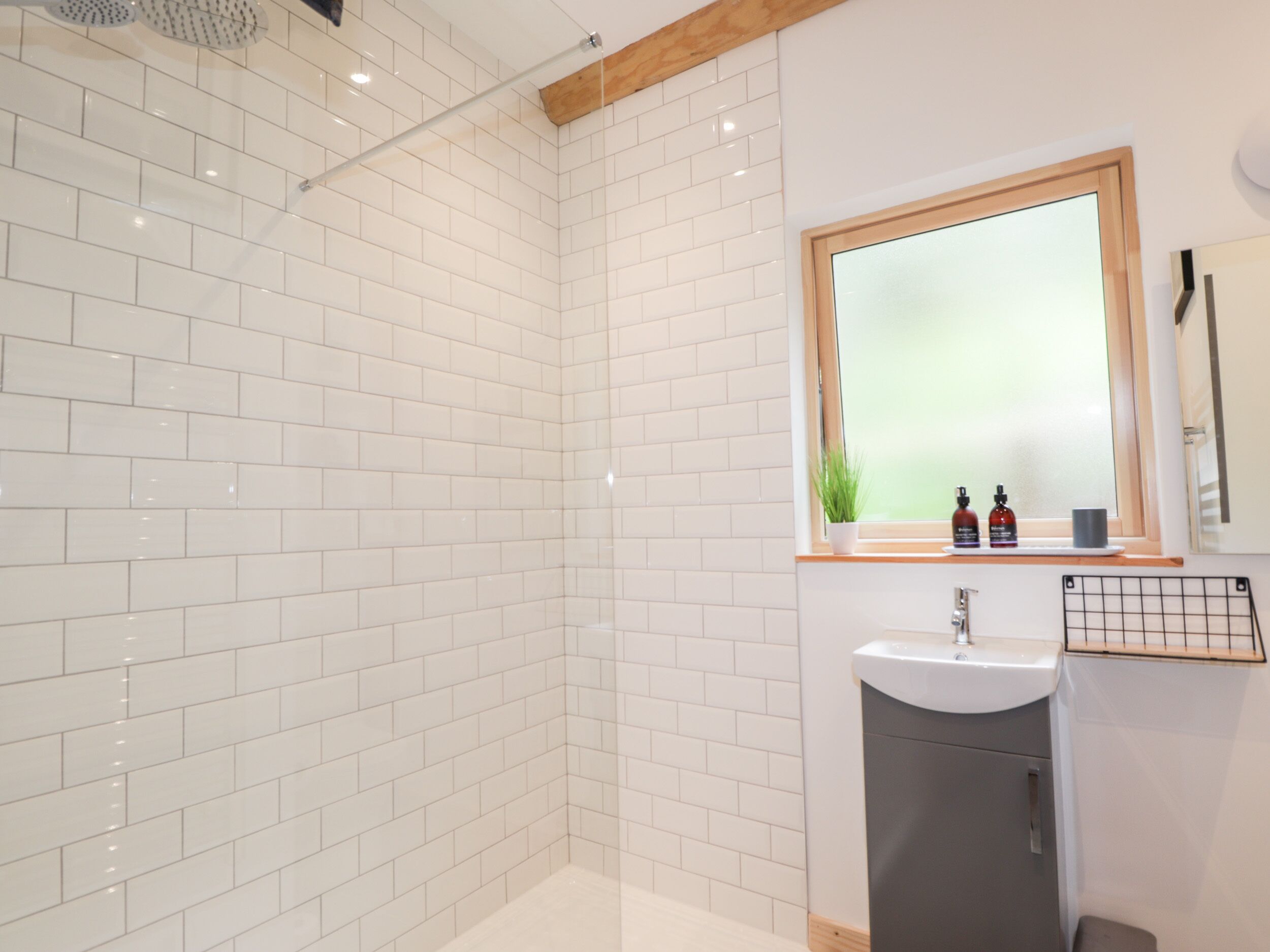Un baño moderno con azulejos blancos tipo metro, techo con vigas de madera, un espejo grande y un lavamanos con un estante pequeño encima.