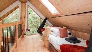 Interior de madera con techo inclinado, una cama con almohadas y una manta, una mesita y un ventanal con vistas a la vegetación.