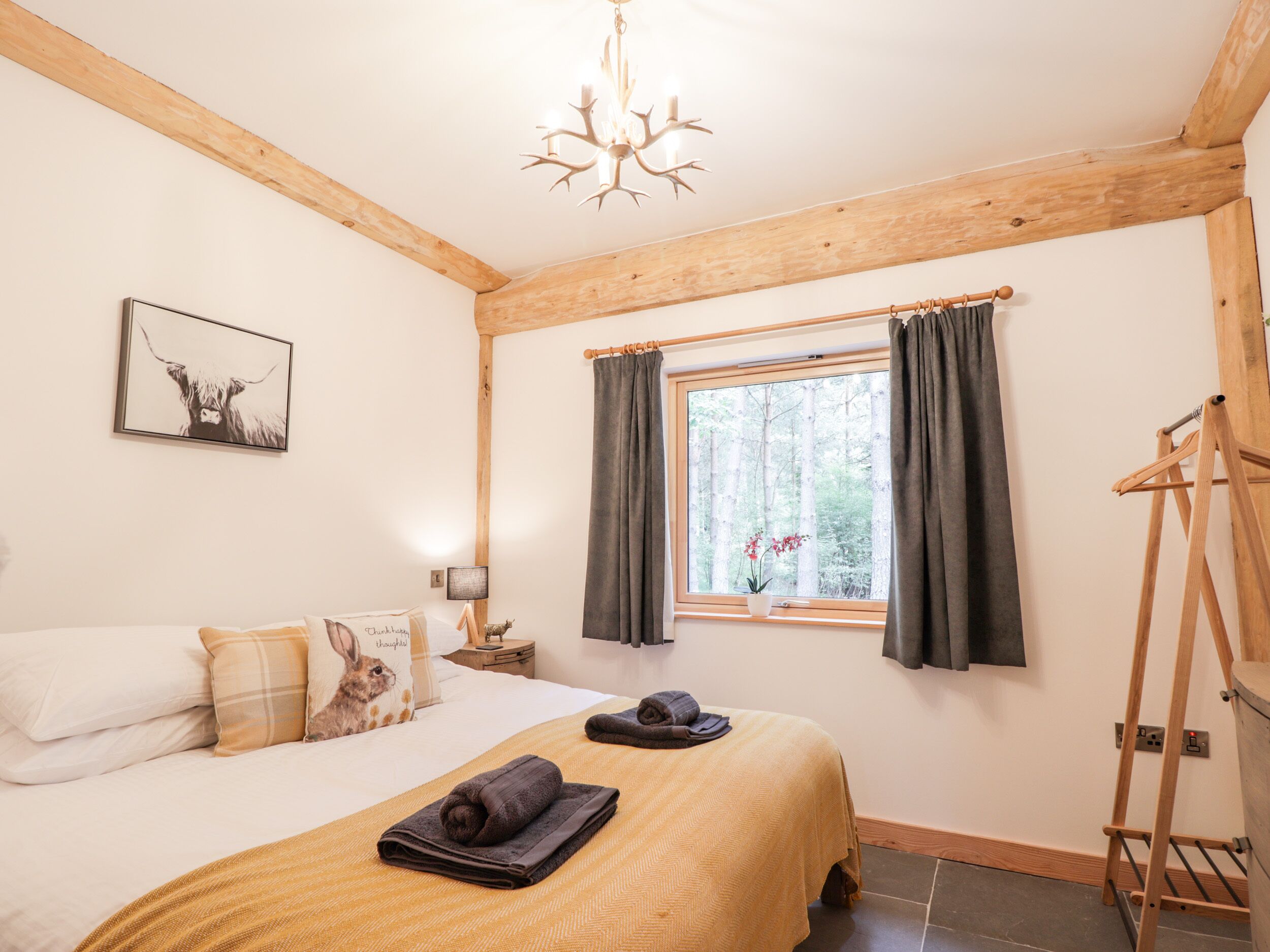 A bedroom with a bed, a window with curtains, a chandelier, a bedside table, and a wall-mounted picture.