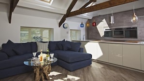 En modern loftlägenhet med synliga träbjälkar, en stor blå soffa i L-form, ett soffbord i glas och ett kök med inbyggda vitvaror.