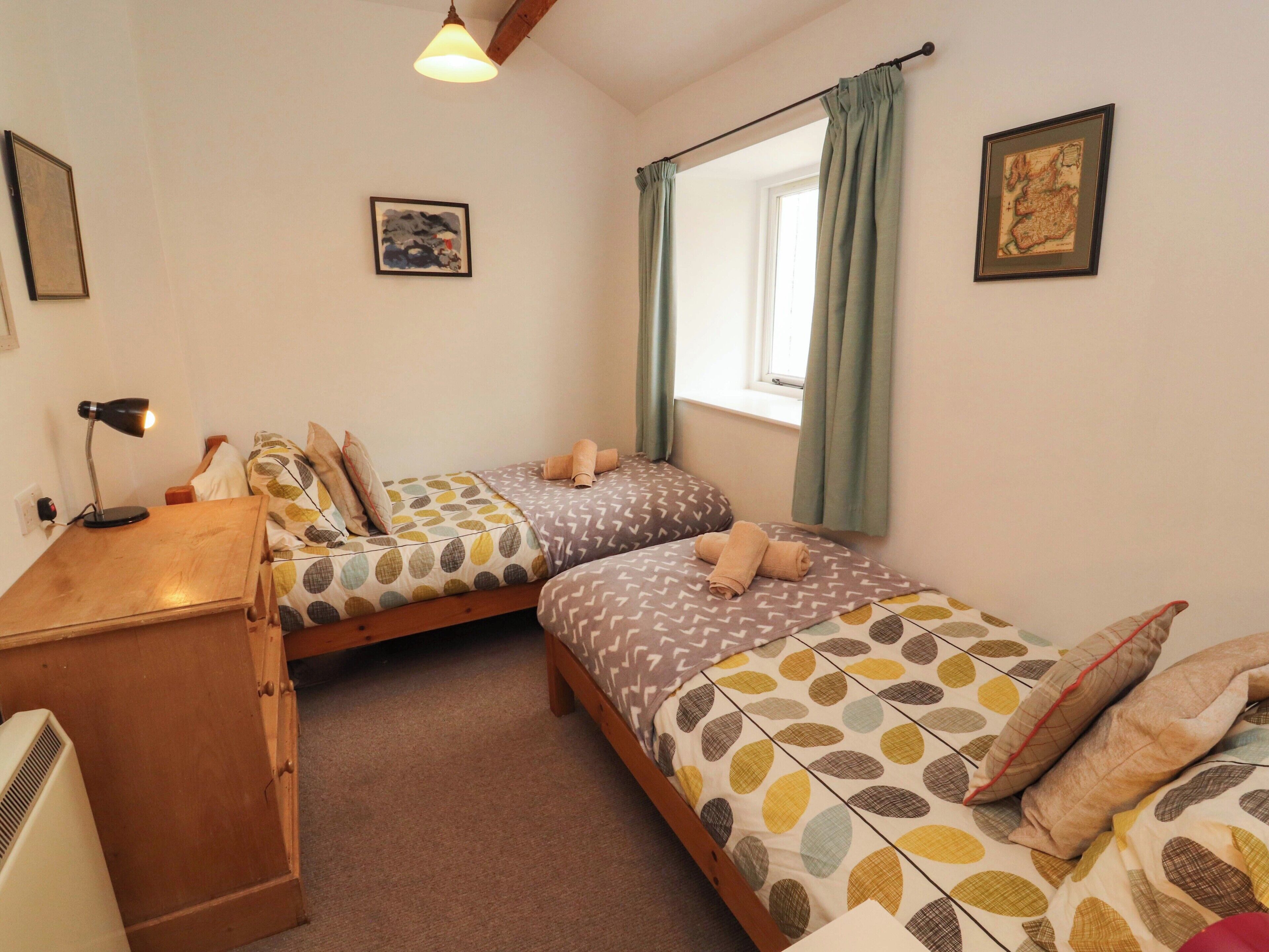 Une chambre avec deux lits, un bureau, une lampe, une fenêtre avec des rideaux et un mur orné d’un tableau encadré.