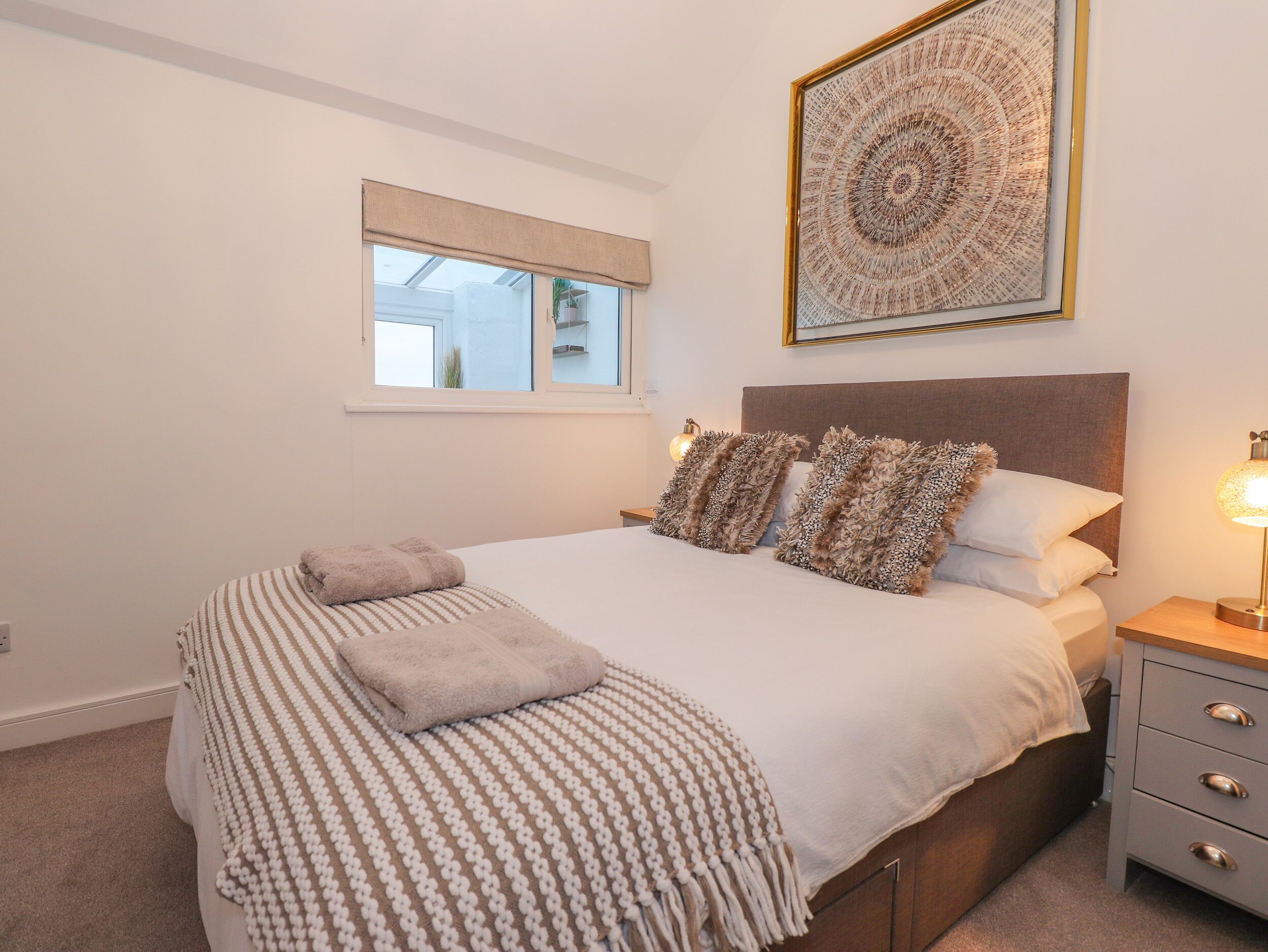 A bedroom with a bed, a bedside table, a lamp, a window, and a framed artwork on the wall.