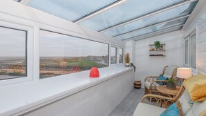 A sunlit room with a view of the beach, a sofa, a chair, and a small table with a lamp. - The Lookout (Millom)