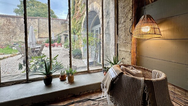 Una habitación rústica con un amplio ventanal que da a un jardín, un sillón de mimbre y una lámpara colgante.