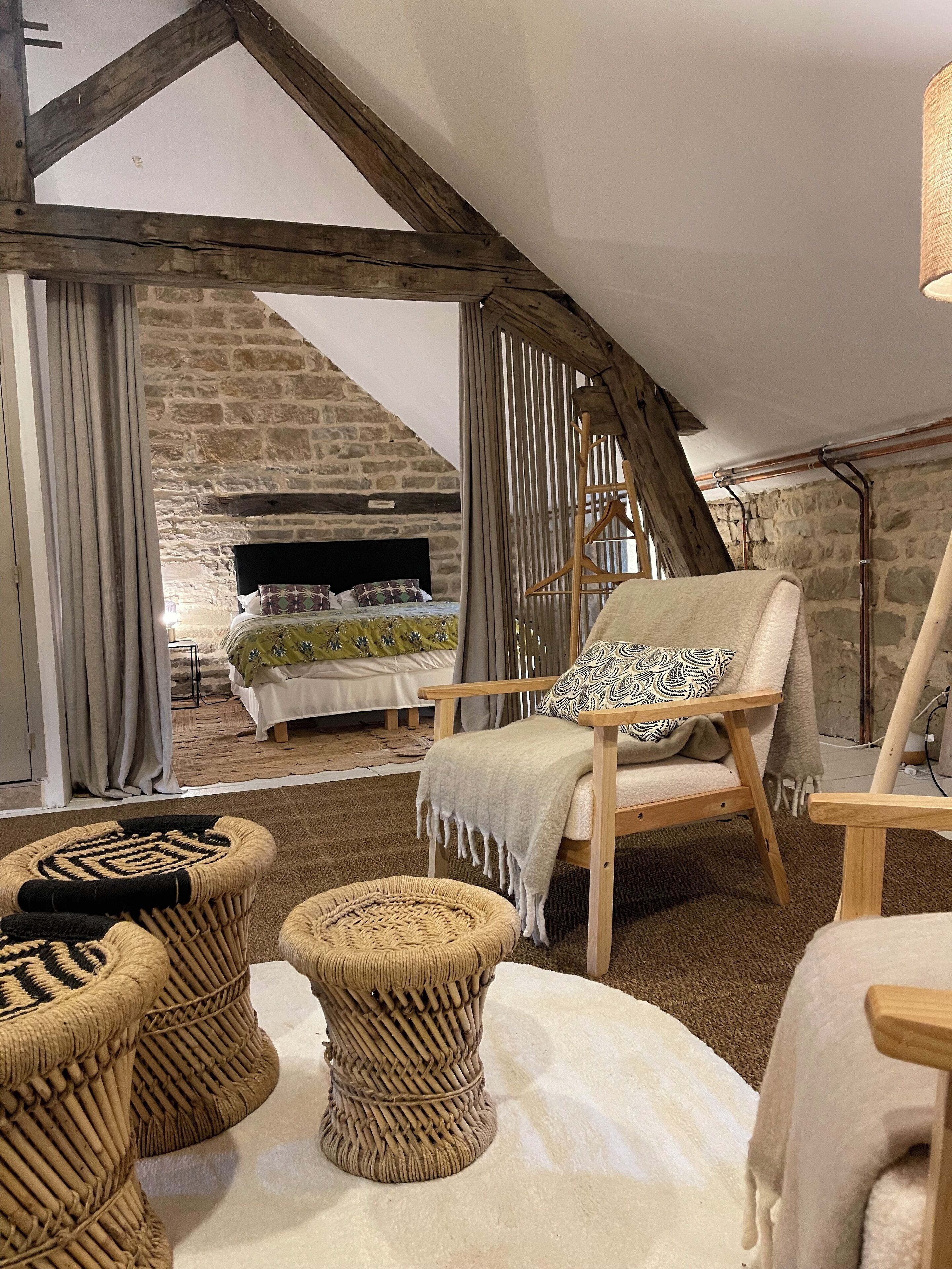 Une pièce chaleureuse avec une cheminée en pierre, des poutres apparentes et un coin salon confortable comprenant un fauteuil et deux tabourets en osier.