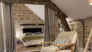 Una habitación acogedora con chimenea de piedra, vigas de madera y una zona de estar cómoda con un sillón y dos taburetes tejidos.