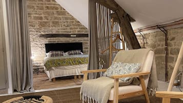 Une pièce chaleureuse avec une cheminée en pierre, des poutres apparentes et un coin salon confortable comprenant un fauteuil et deux tabourets en osier.