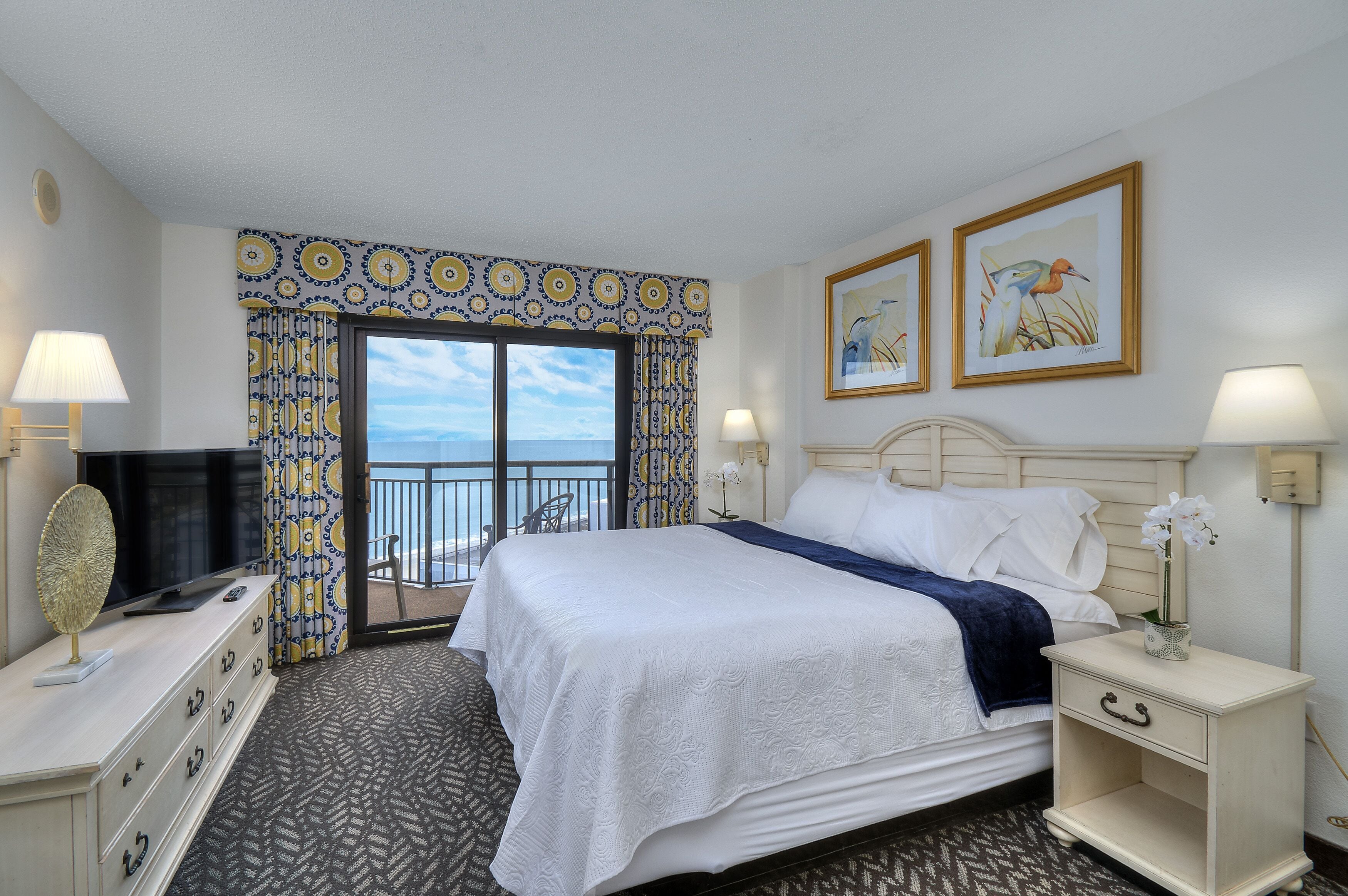A hotel room with a bed, a TV, a dresser, and a balcony with a view.
