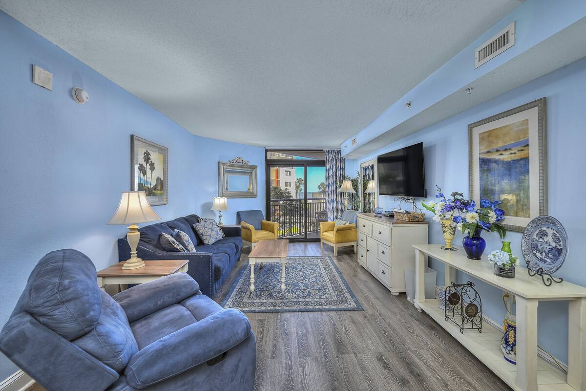 A living room with a blue sofa, yellow chairs, a wooden coffee table, and a TV on a white stand.