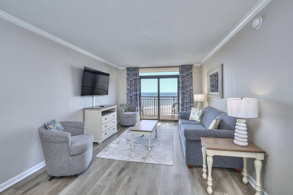 A living room with a grey armchair, a sofa, a coffee table, a TV mounted on the wall, and a balcony with a view.