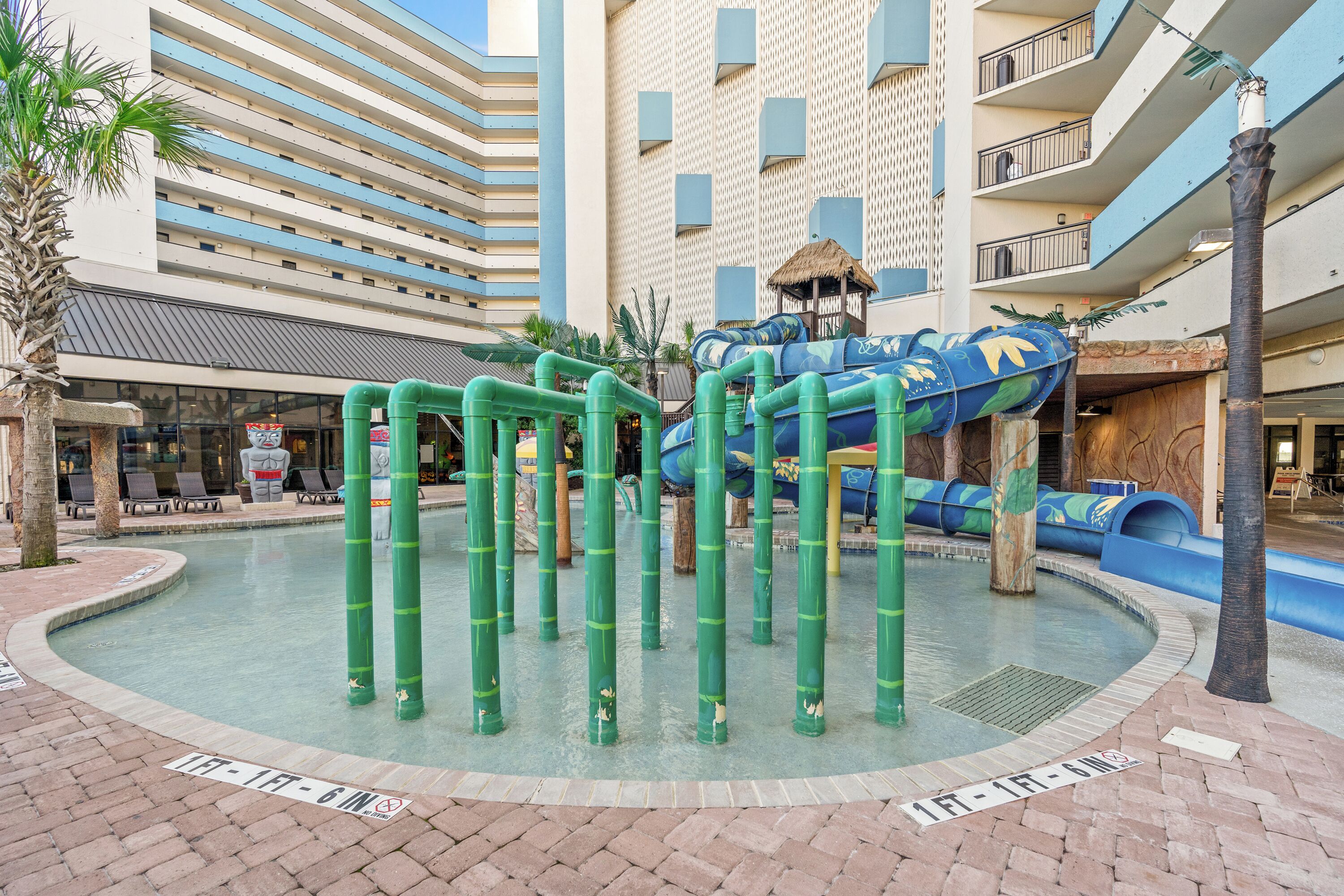 A water play area with slides and a pool, surrounded by a brick walkway and palm trees.