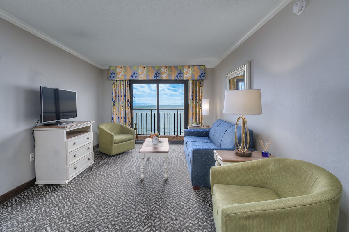A hotel room with a television, a sofa, a chair, a small table, and a view of the balcony.