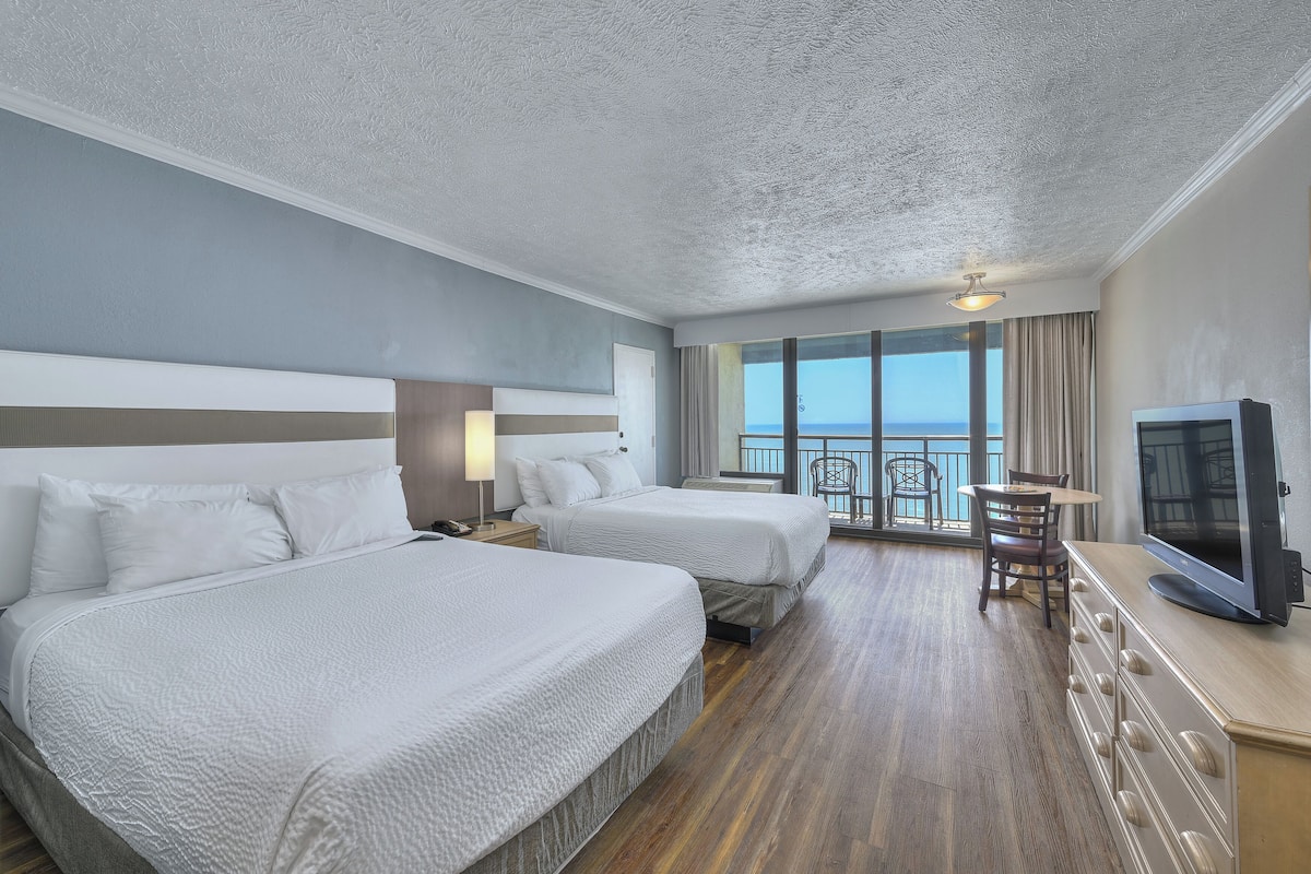 A hotel room with two beds, a TV, a dining area with a table and chairs, and a balcony with a view.