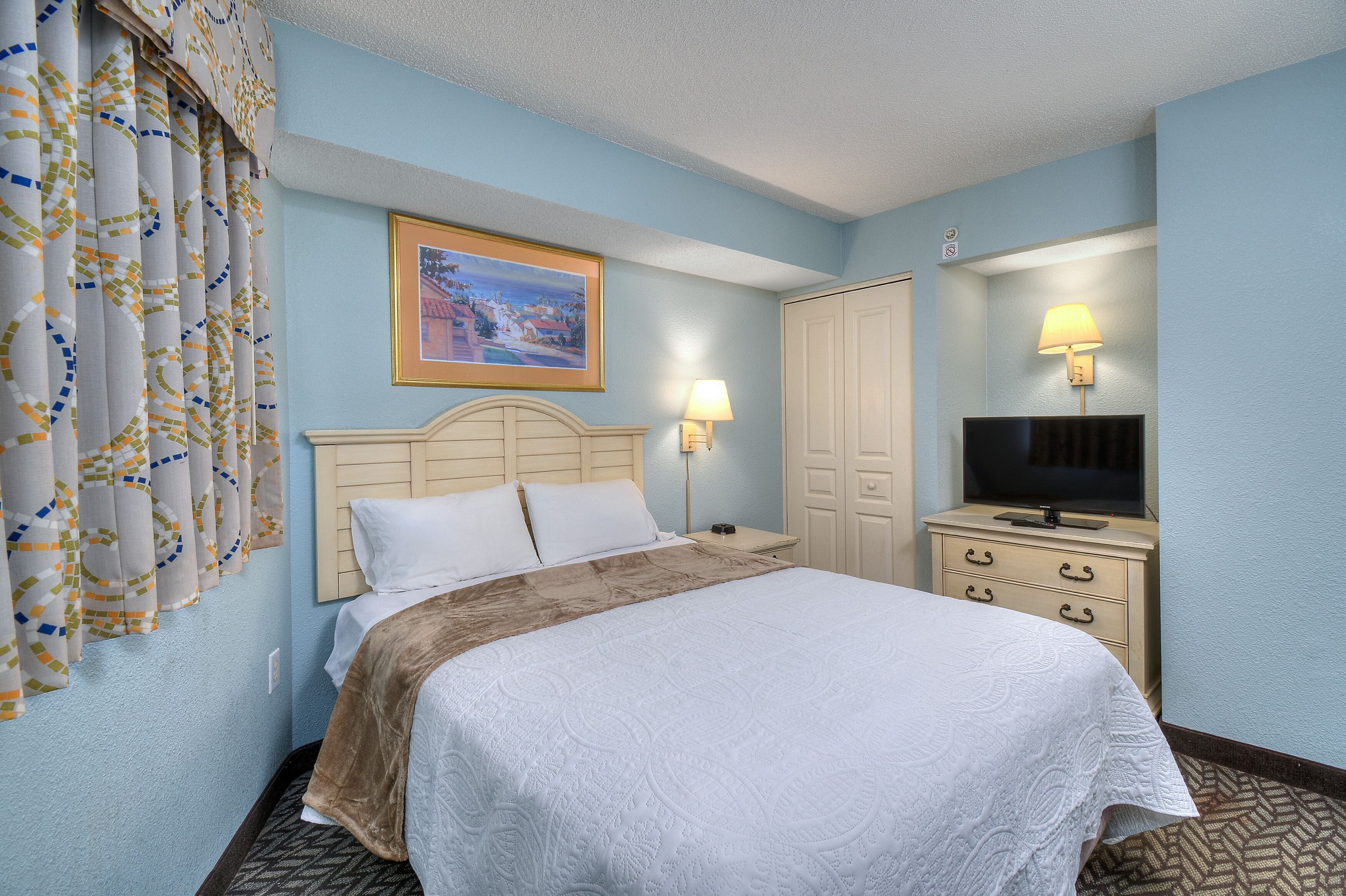 A hotel room with a bed, a television, a dresser, and a painting on the wall.