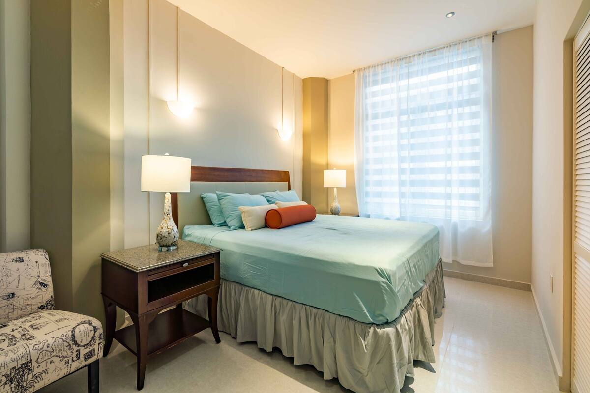 Una camera d'albergo con un letto, lampade sui comodini, un tavolino, una sedia e un'ampia finestra con tende.
