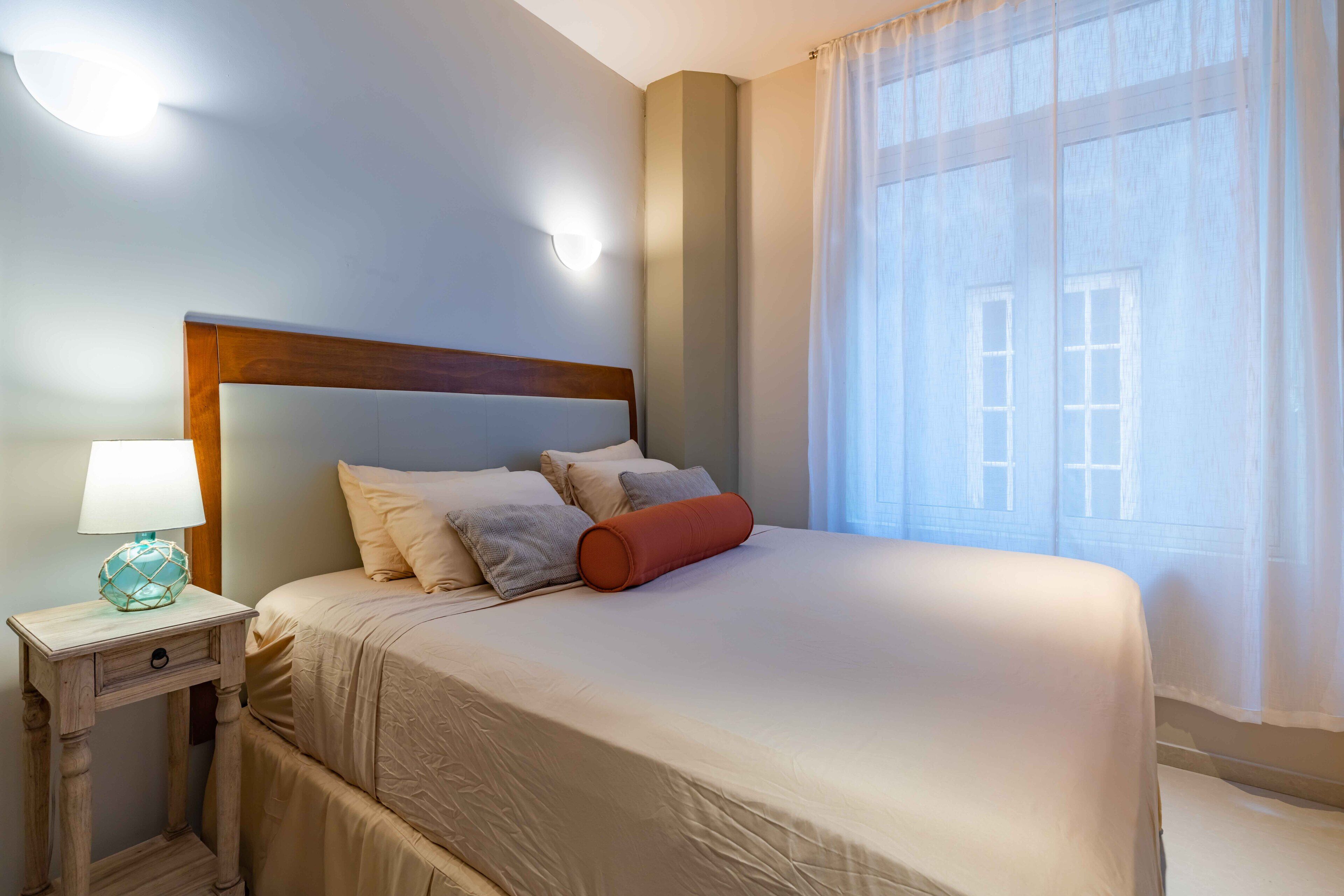 A neatly made bed with a wooden headboard, a bedside lamp, and a window with sheer curtains.