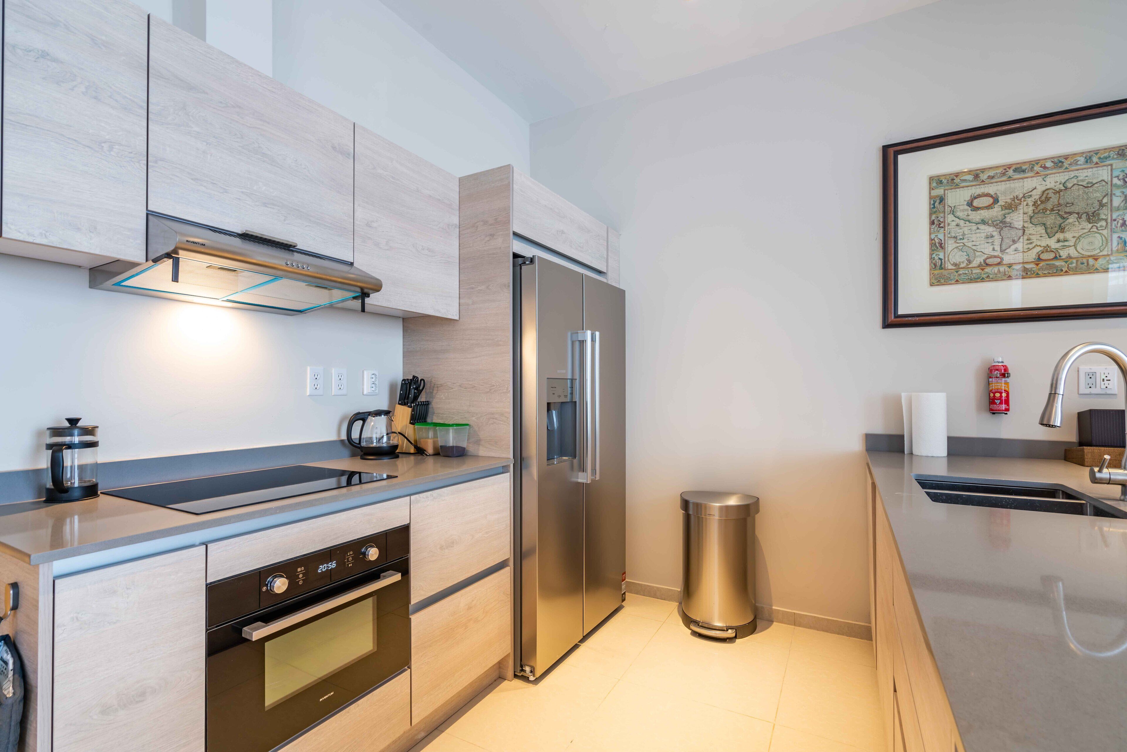A modern kitchen with light wood cabinets, stainless steel appliances, a built-in oven, and a stainless steel refrigerator.