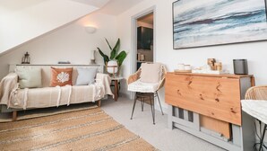 A cozy loft bedroom with a sloped ceiling, a large window, a bed, a wooden dresser, a chair, and a painting on the wall.
