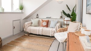 A cozy living room with a high ceiling, a sloped ceiling, a sofa with cushions, a wicker chair, a wooden table, and a large plant.
